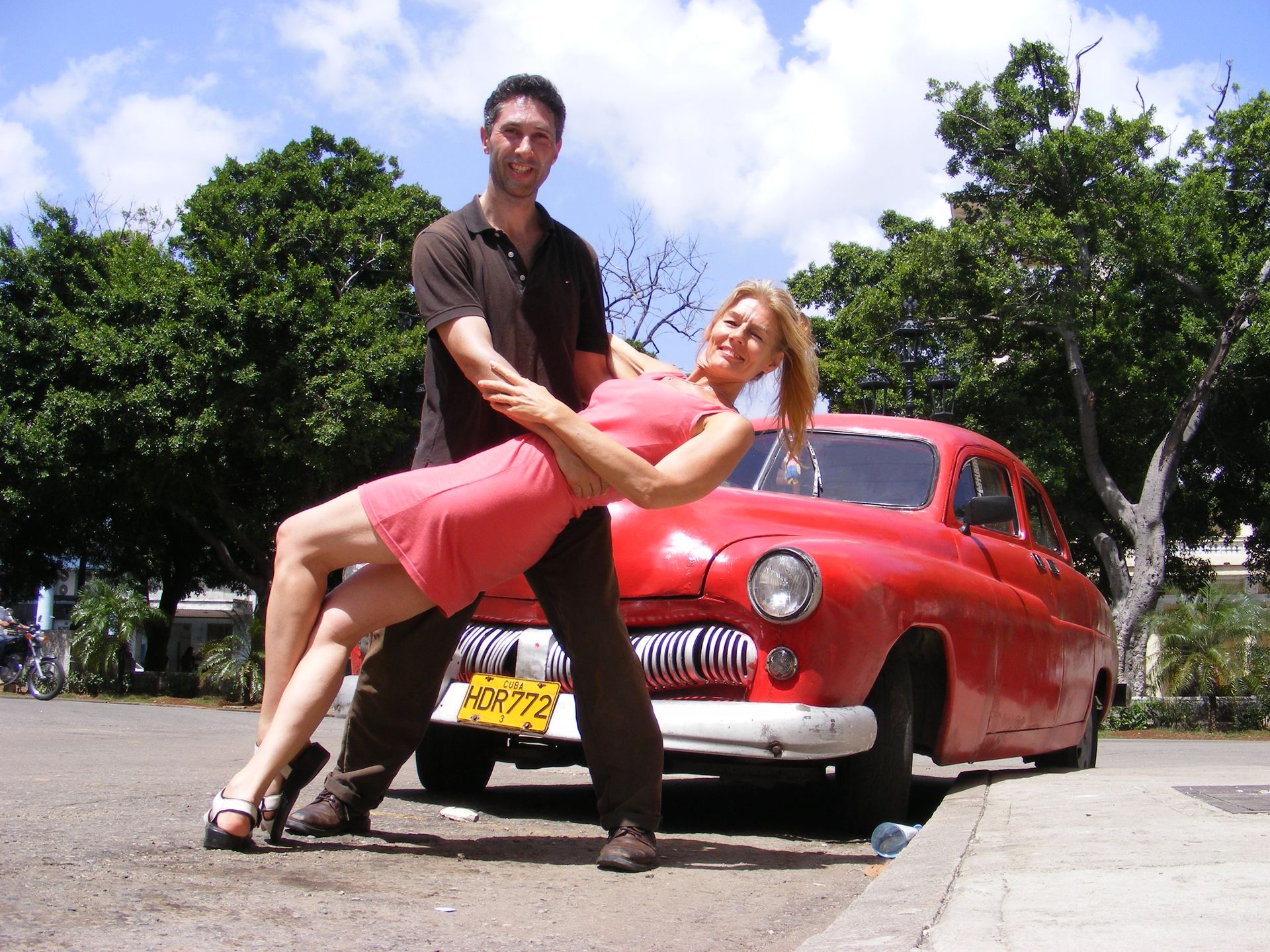A salsa couple holds a dip beside a classic red car on a sunny Havana street